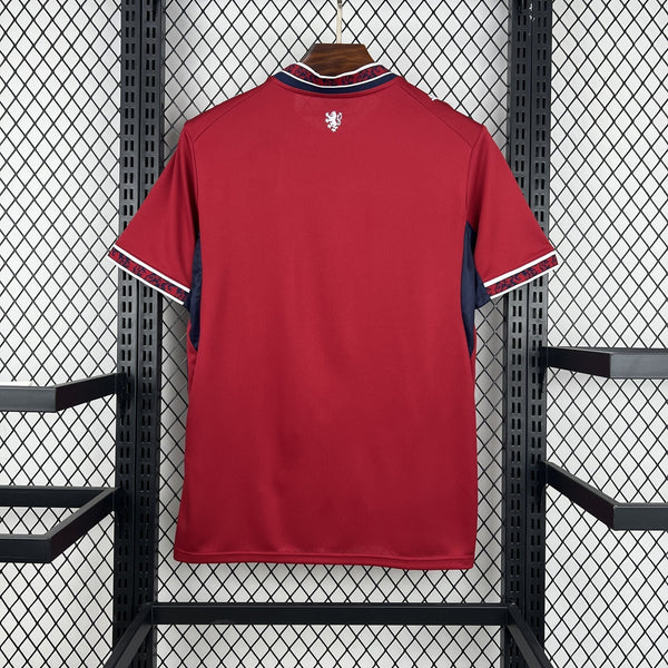 Red sports jersey with a logo on the chest, hanging against a black metal fence.